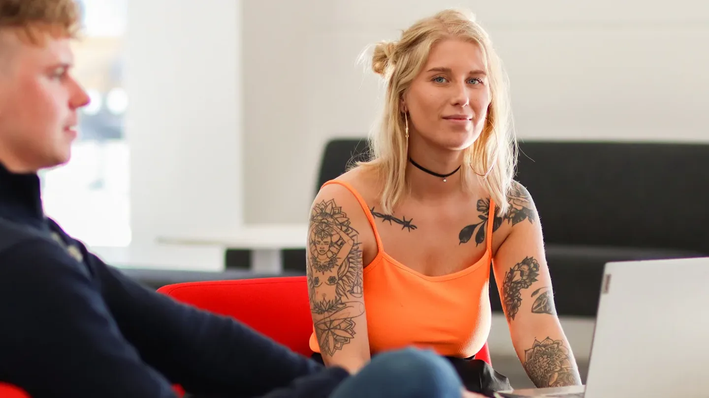 Ung leende kvinna med tatueringar som tittar in i kameran. Till vänster om henne sitter en ung man.
