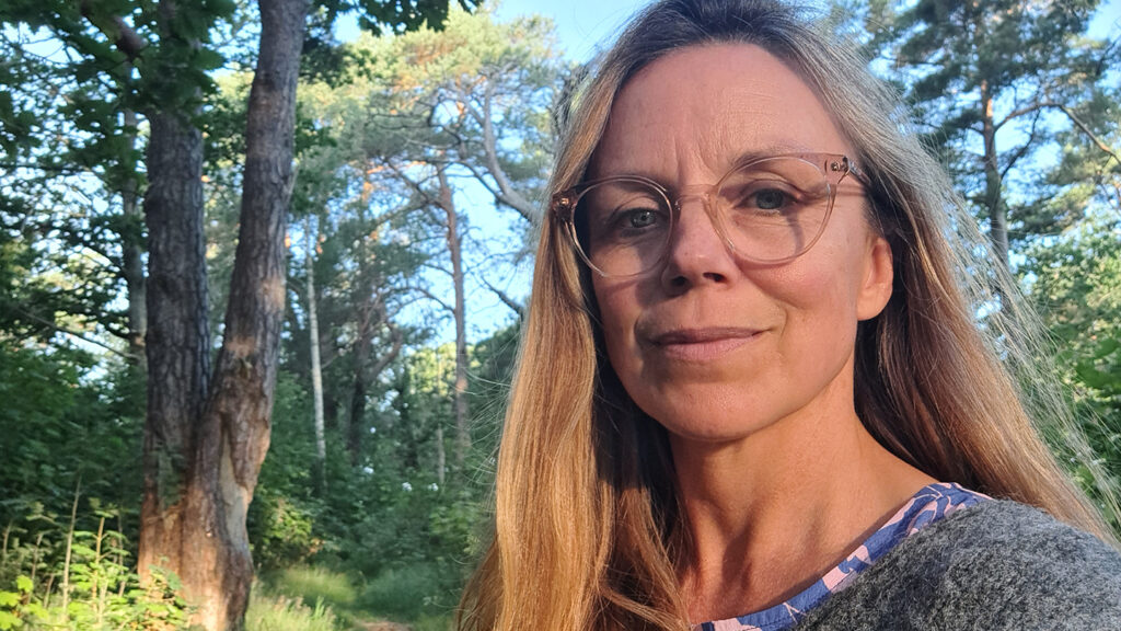 Specialistundersköterskan Josefine Lindvall står i skogsmiljö och tittar in i kameran. Josefine syns i högra sidan av bilden och i bakgrunden, till vänster syns ett lövträd.