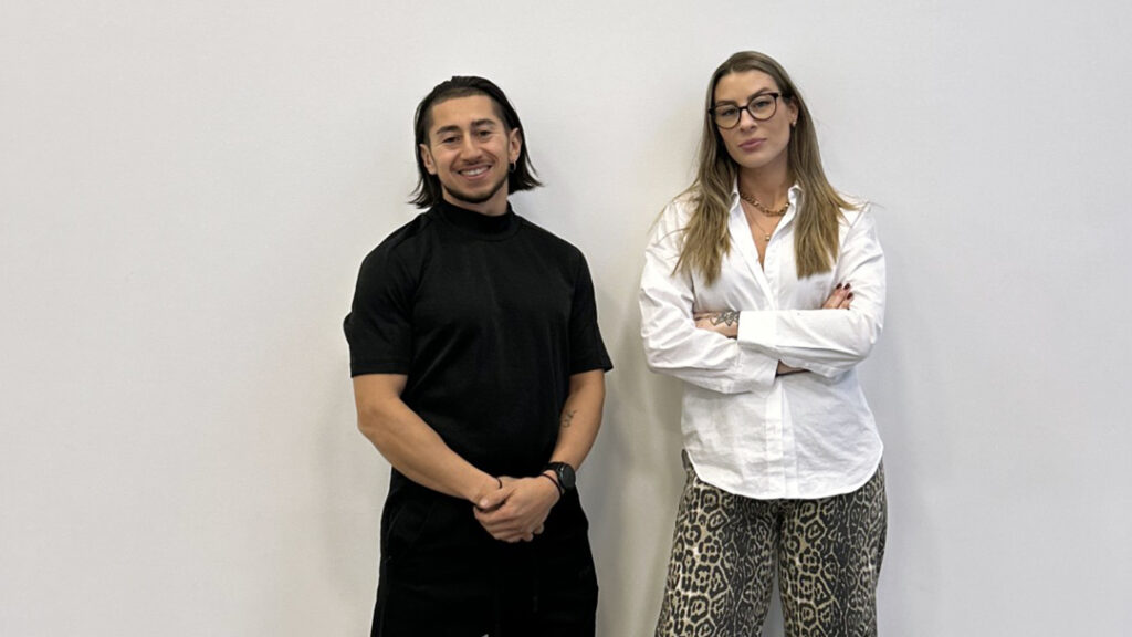 Johannes Ojanperä och Amanda Sörensen poserar framför en vit vägg. Johannes har mörkt axellångt hår och klädd helt i svart. Amanda har långt, brunt hår, bär mörka glasögon och en vit skjorta med mörka byxor.
